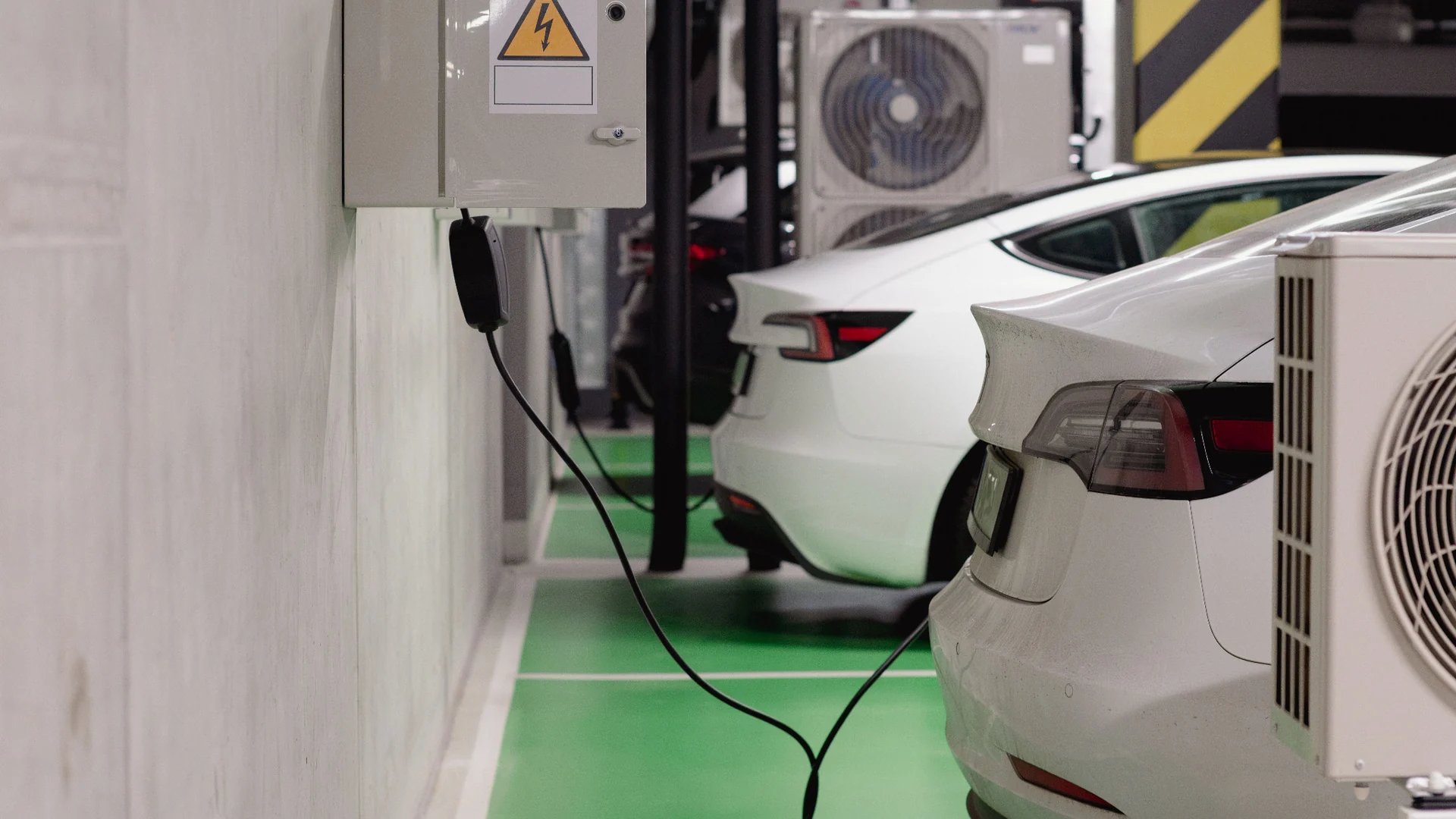 Two electric cars charging from a wall-mounted unit in an indoor parking bay